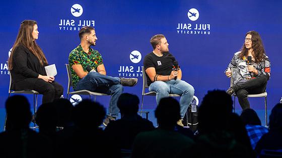 Four people seated on a stage, talking and holding microphones in front of a large crowd.